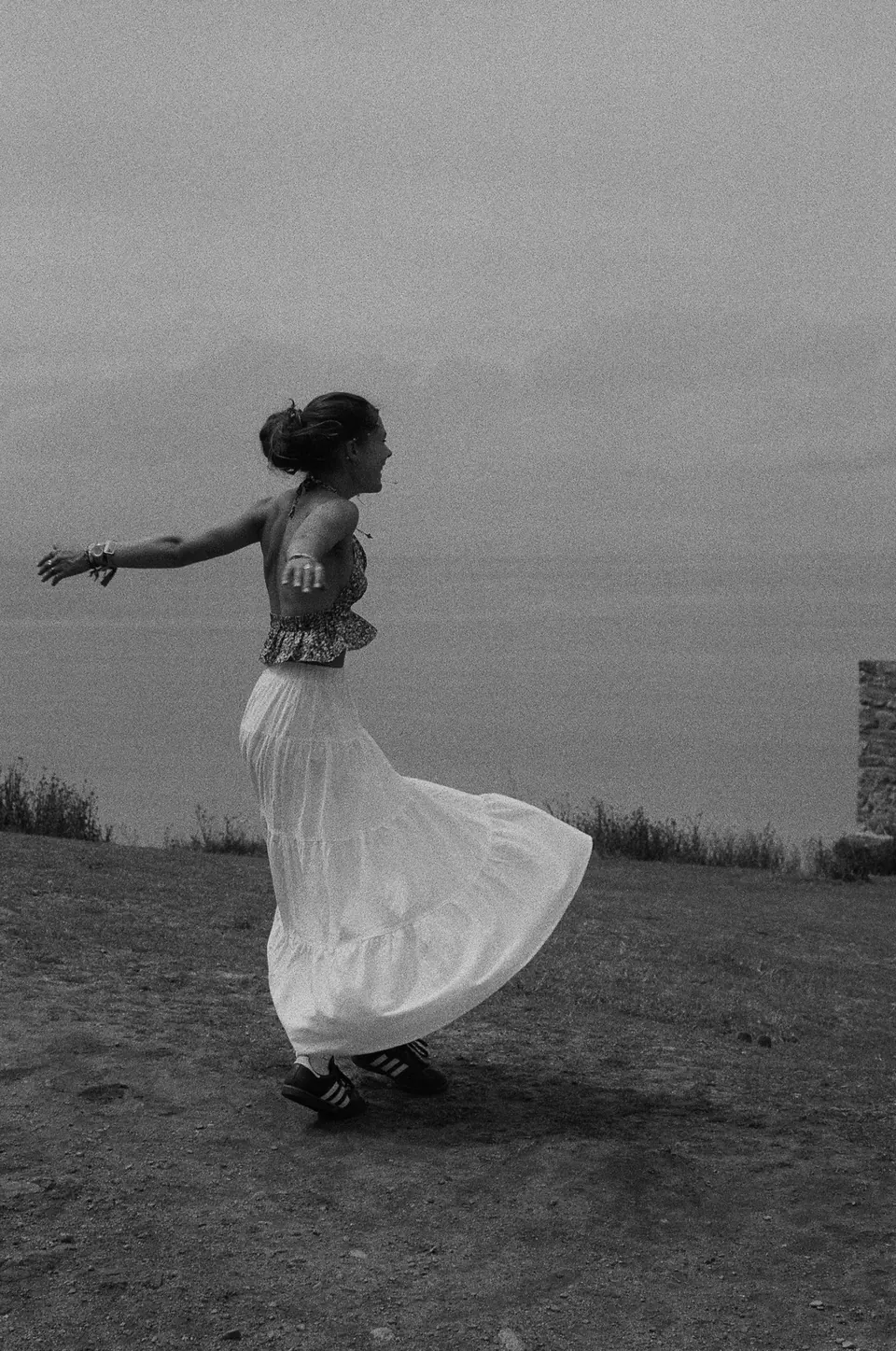 Photographie en noir et blanc : une femme en jupe longue blanche tourne sur elle-même, les bras écartés, face à la mer.