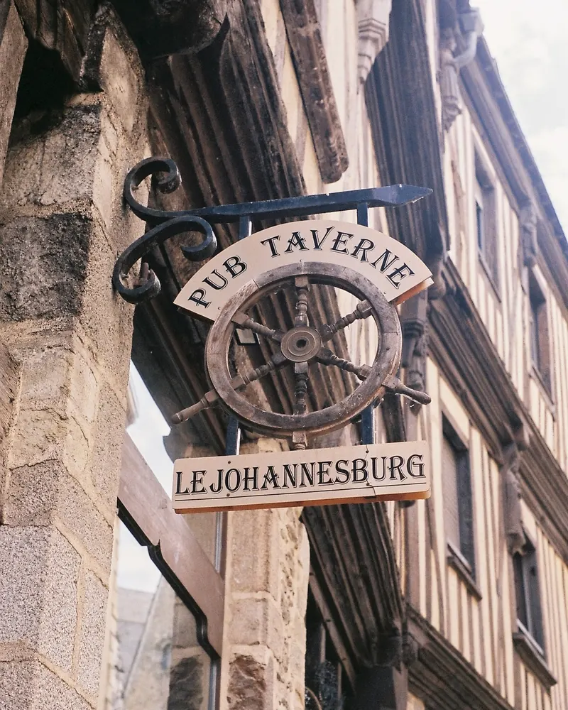 Une enseigne en fer forgé en forme de barre à roue, scellée à une façade de pierre, portant les mots « Pub Taverne Le Johannesburg ».