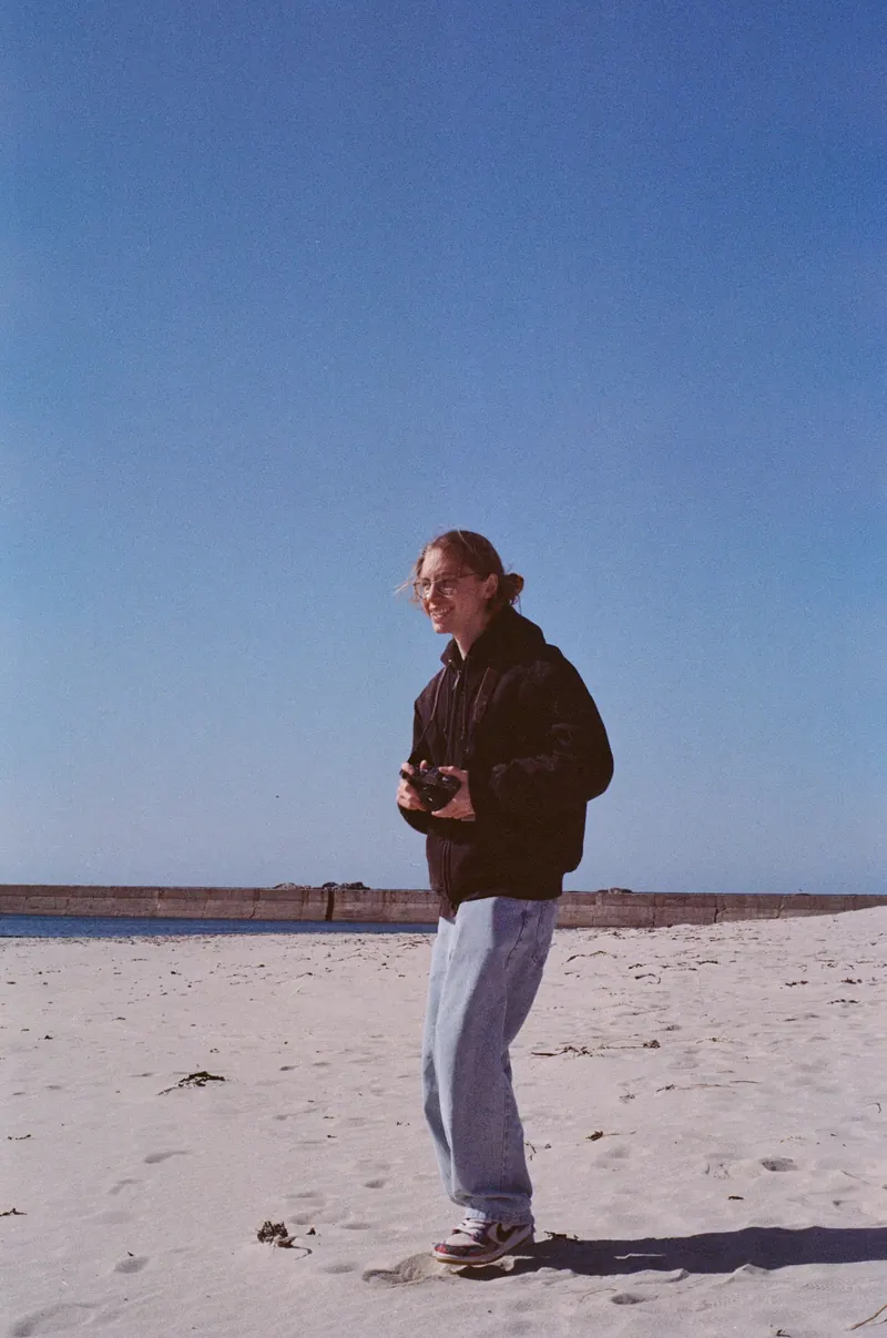 Un homme en veste sombre et jean clair marche sur une plage de sable, un appareil photo devant lui, devant une longue digue de pierre.