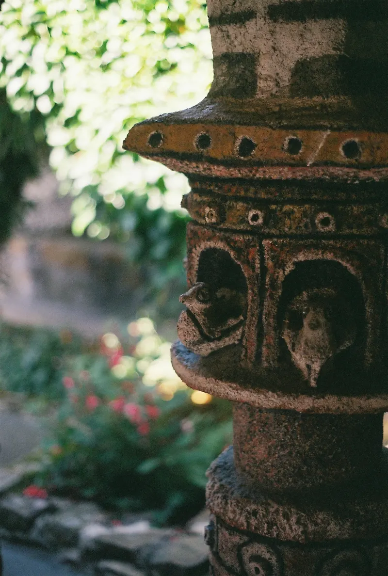 Une lanterne de pierre sculptée, de petites figures logées dans ses niches, devant un jardin flou où percent des fleurs rouges et des feuillages au soleil.