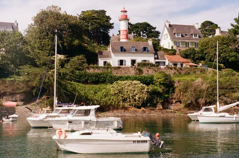 Un phare blanc à toit rouge domine une anse où mouillent quelques voiliers et vedettes, devant des maisons à toit d’ardoise.