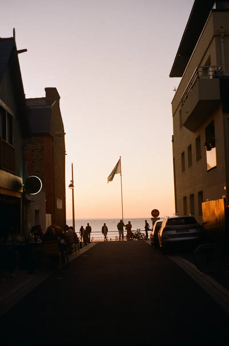 Une rue en pente descend vers la mer au couchant ; des passants sont attablés en terrasse et des silhouettes se découpent à contre-jour.