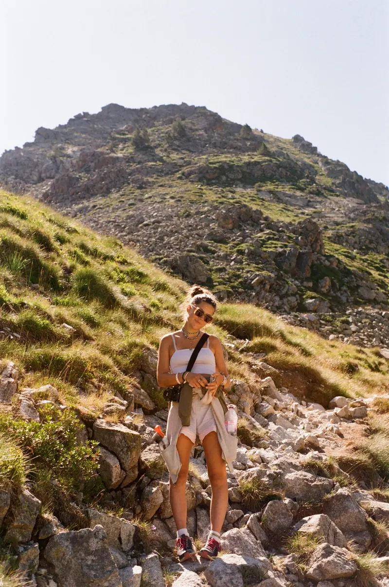 Une jeune femme en short et débardeur clair monte un sentier caillouteux, un appareil photo à la main, entre les herbes hautes et les rochers d’un versant de montagne.