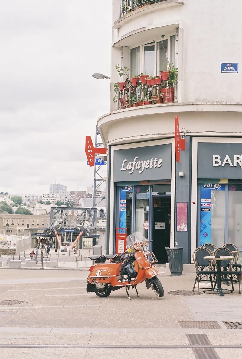 Un scooter Vespa orange garé sur une place devant le café-tabac « Lafayette », à l’angle d’un immeuble blanc aux balcons fleuris ; au fond, les pylônes rouges d’un téléphérique et la ville.