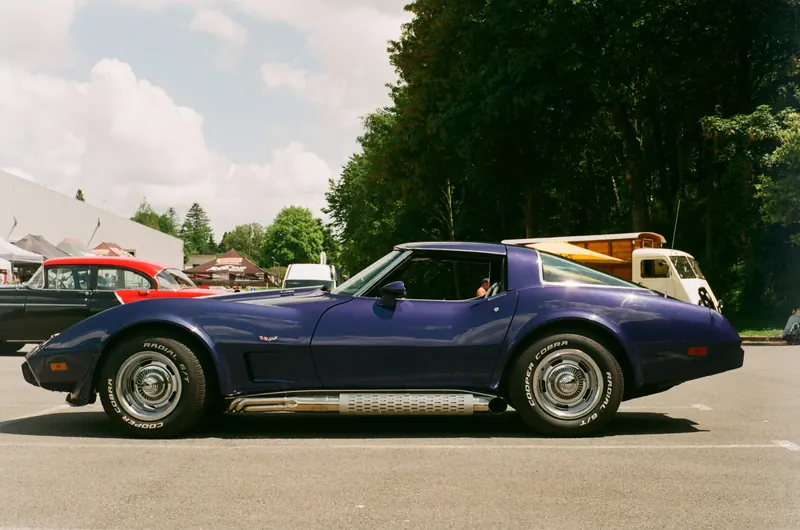 Une Corvette C3 violette photographiée de profil sur un parking, devant une rangée d’arbres et d’autres voitures anciennes.