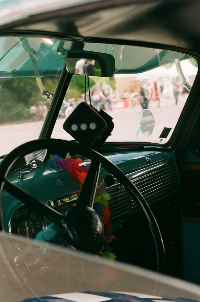 L’habitacle d’une voiture ancienne vu par la vitre : volant à jante fine, compteur rond, un désodorisant suspendu au rétroviseur et un collier de fleurs posé sur la colonne de direction ; par le pare-brise, la foule floue d’un rassemblement automobile.