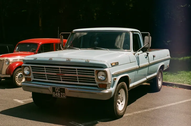 Un pick-up Ford F-100 bleu clair de trois quarts avant sur un parking, un utilitaire ancien rouge et crème garé derrière.