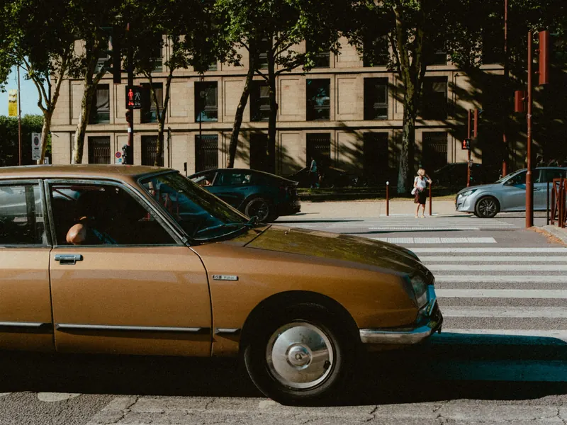 Une Citroën ancienne de couleur ocre à l’arrêt devant un passage piéton, de profil, l’ombre des arbres posée sur sa carrosserie.