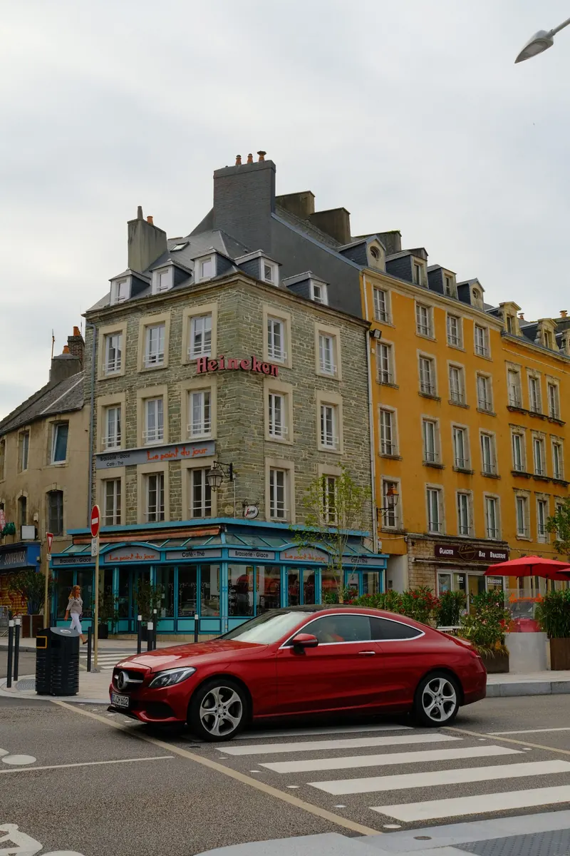 Un coupé rouge à l’arrêt devant un carrefour, au pied d’un immeuble de granit et d’une façade jaune.