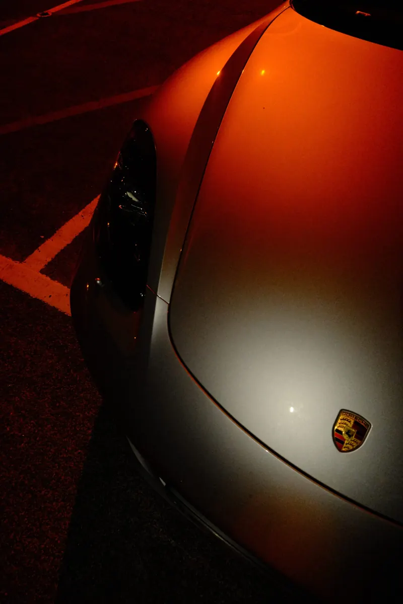 Détail de l’aile avant d’une Porsche argent la nuit : l’écusson, le bord du phare et la courbe de l’aile se détachent dans la lumière orange.