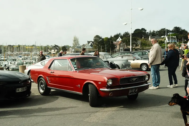Une Ford Mustang ancienne rouge, de trois quarts avant, à un rassemblement de voitures sur le parking d’un port ; des visiteurs l’entourent, des voiliers au fond.