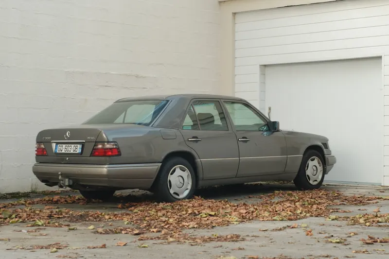 Une Mercedes W124 grise vue de trois quarts arrière devant la porte blanche d’un garage, le sol couvert de feuilles mortes.