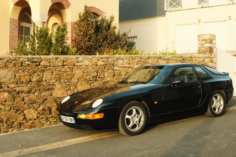 Une Porsche noire garée de trois quarts avant le long d’un muret de pierre, la lumière rasante de fin de journée glissant sur l’aile et le capot.