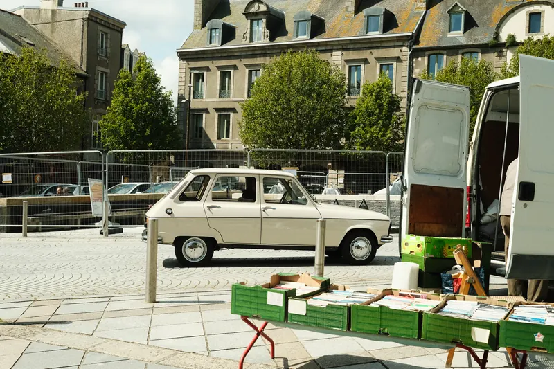Une voiture ancienne blanche garée de profil sur une place pavée, devant les façades de granit d’un jour de marché.