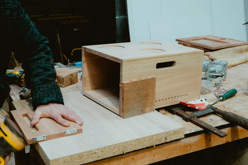 Caisson de borne en bois, percé d’ouvertures rondes, en cours d’assemblage sur un établi, à côté d’un serre-joint ; une main tient une petite pièce de bois équipée de charnières.