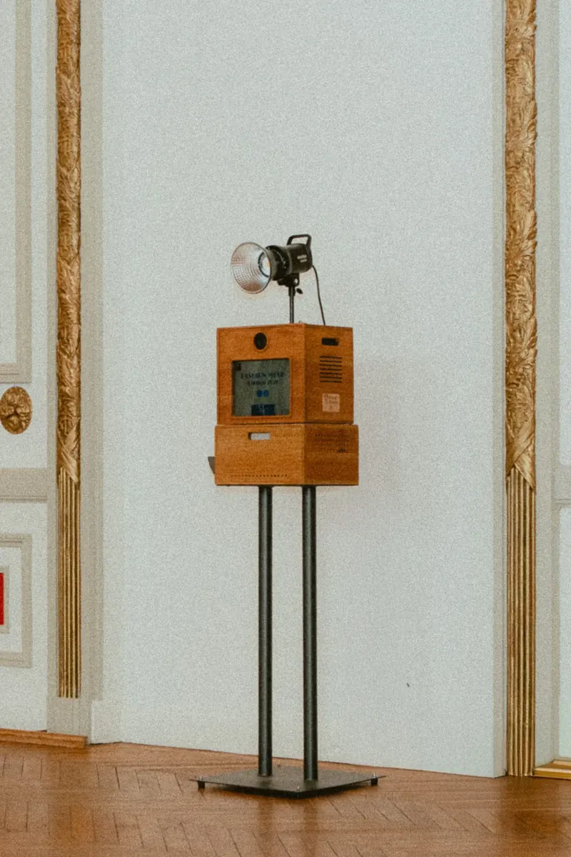 Borne photo en bois à deux caissons, écran allumé, flash de studio fixé au sommet, entre deux pilastres dorés d’un salon blanc.
