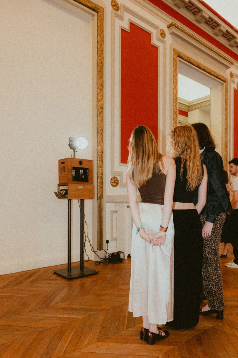 Trois invités, vus de dos, attendent devant une borne photo en bois installée contre le mur d’un salon aux panneaux rouges et aux moulures dorées.