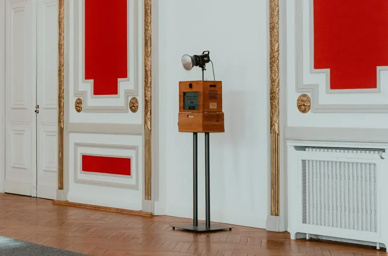 Borne photo en bois posée sur deux pieds métalliques, surmontée d’un flash à réflecteur, contre le mur blanc d’un salon aux panneaux rouges et aux moulures dorées, sur un parquet en chevrons.