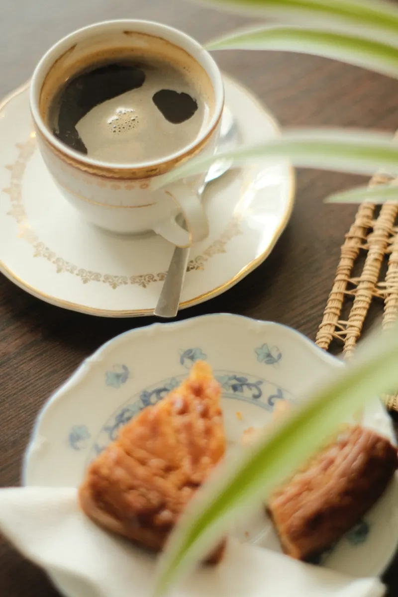 Une tasse de café et deux gâteaux secs posés sur une table en bois, à côté d’une feuille verte, en lumière douce.