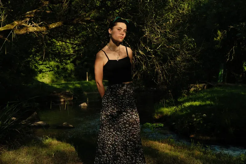 Une jeune femme en débardeur noir et jupe à fleurs, cadrée à mi-corps, éclairée par un rai de soleil devant un ruisseau et un sous-bois sombre.