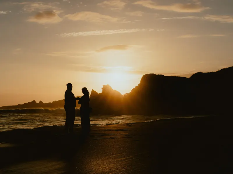 Un couple en silhouette, face à face sur la plage, devant des rochers et le soleil couchant.