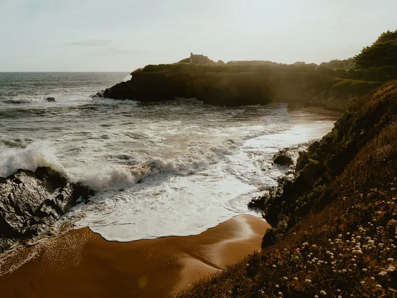 Une crique du Finistère battue par la houle : les vagues déferlent sur le sable, au pied d’une pointe rocheuse.