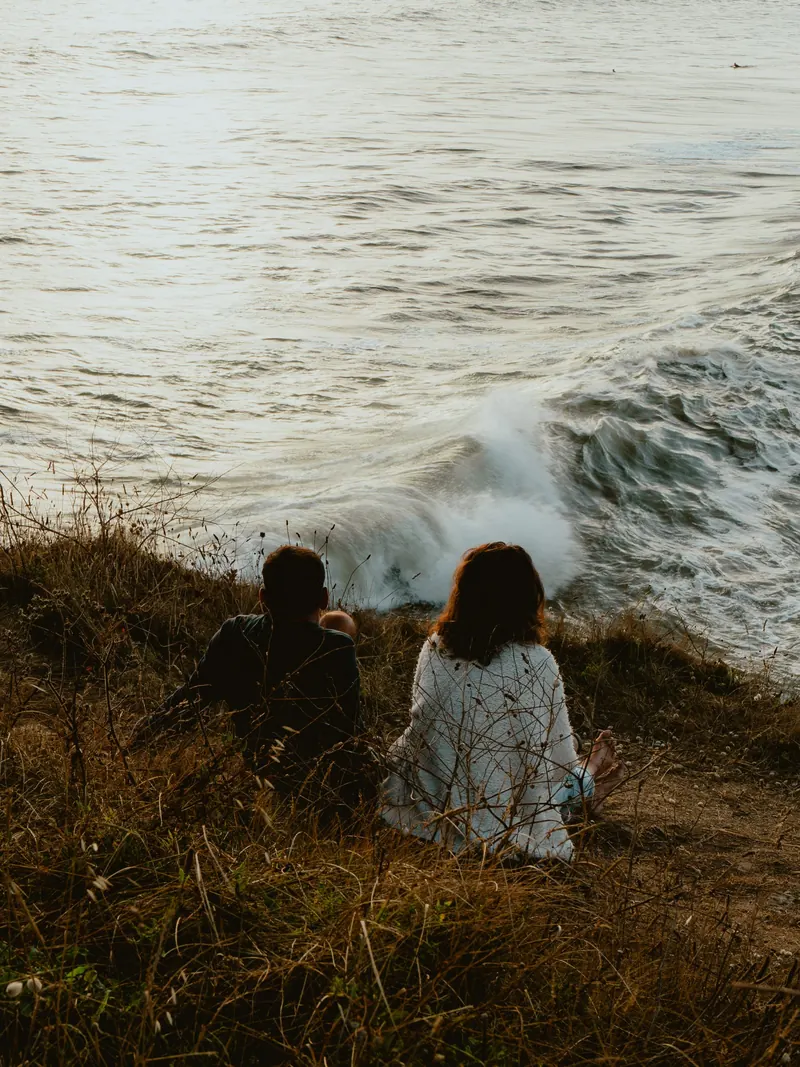 Deux personnes assises de dos dans les herbes sèches regardent les vagues se briser en contrebas, un jour de houle dans le Finistère.