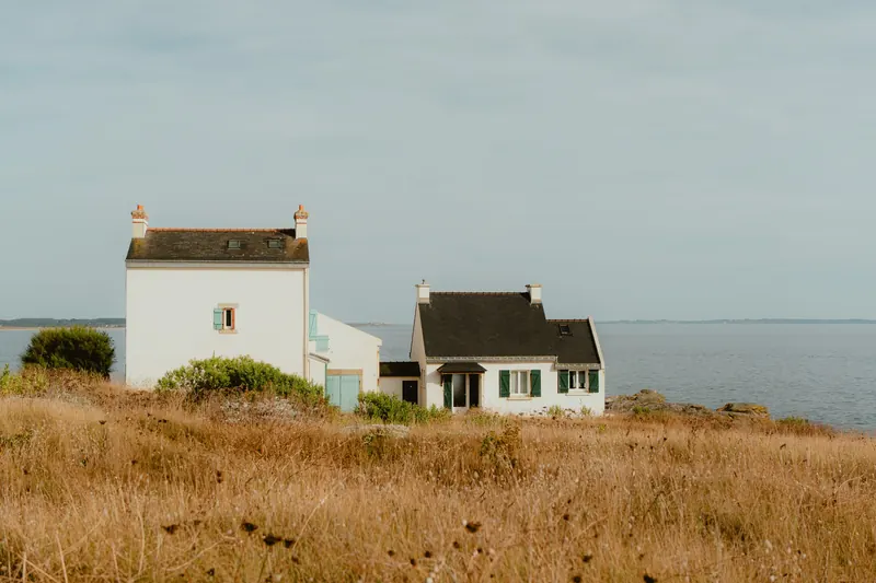 Deux maisons blanches aux toits d’ardoise sur une lande d’herbes sèches, face à une mer calme sous un ciel voilé.