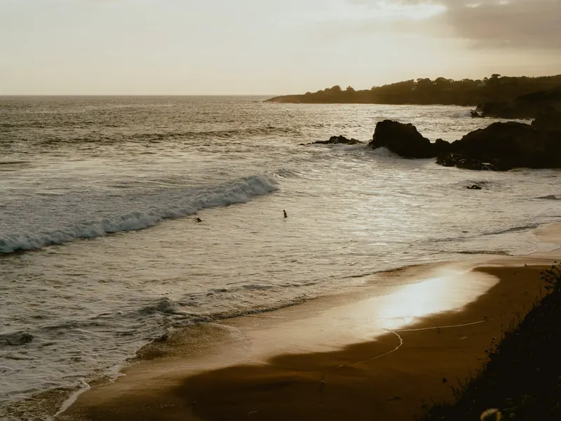 Une plage dans la lumière du soir : les vagues arrivent en biais sur le sable mouillé, près de rochers, et deux personnes sont à l’eau au large.