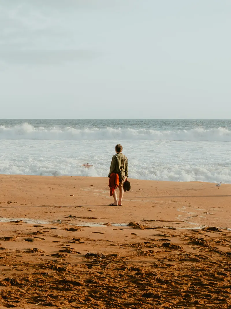 Une personne de dos, pieds nus, un vêtement rouge et ses chaussures à la main, s’arrête sur le sable face aux rouleaux d’écume.