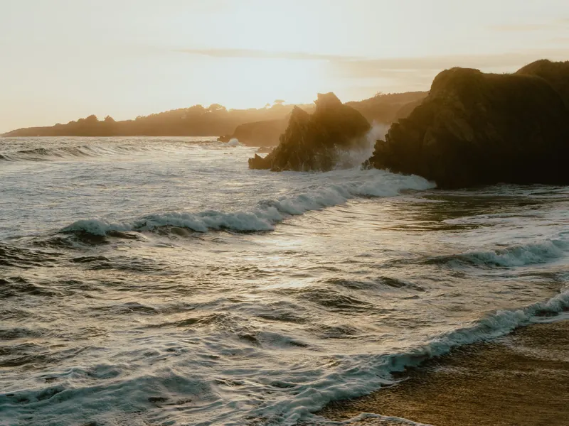 Des rochers à contre-jour dans la lumière dorée du soir ; la houle s’étale en rouleaux jusqu’à la plage.