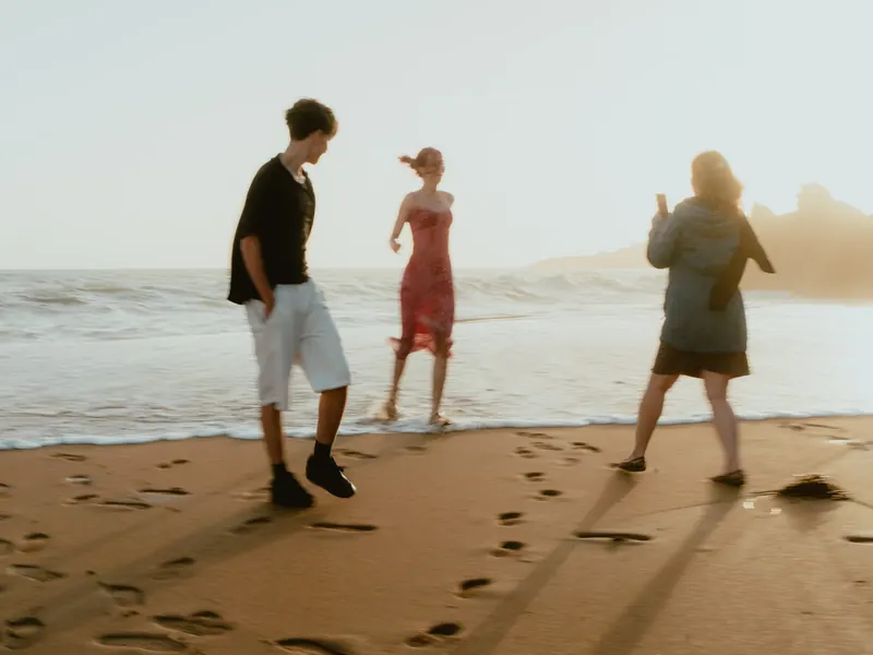 Sur une plage à contre-jour, un jeune homme marche sur le sable, une jeune femme en robe rouge avance au bord de l’eau et une femme tient son téléphone levé.