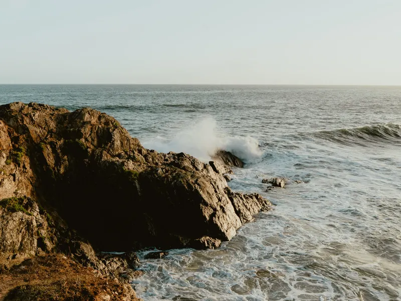 Une vague se brise contre un gros rocher au bord de la mer, dans la lumière de fin de journée.