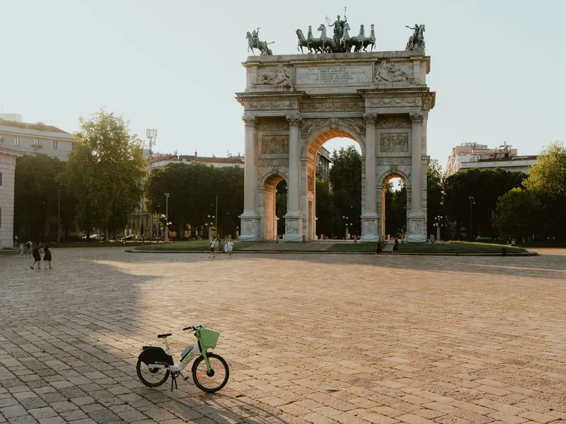 L’Arc de la Paix, surmonté d’un char et de statues équestres, au bout d’une vaste place pavée ; au premier plan, un vélo vert à panier, seul, dans une lumière dorée.