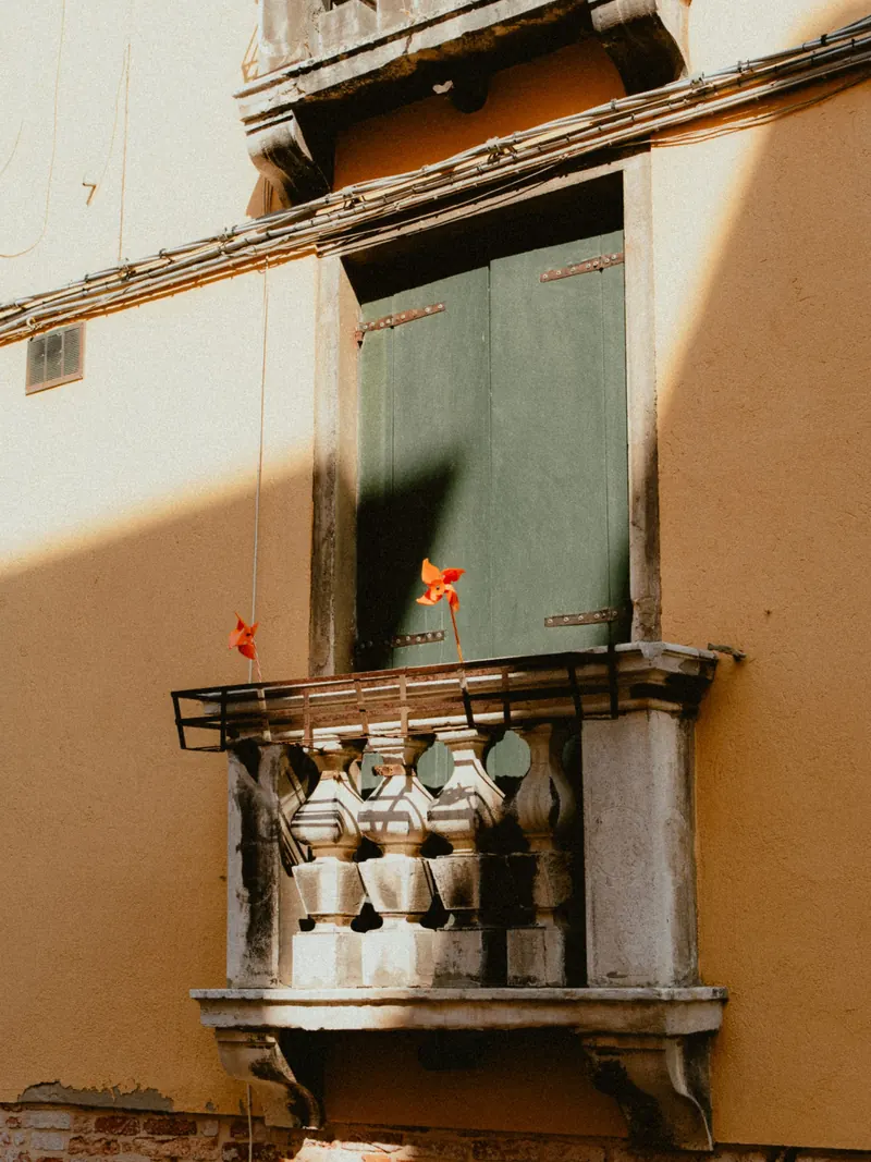 Deux moulinets orange piqués dans la rambarde d’un balcon de pierre à balustres, devant des volets verts et un mur ocre coupé par une ombre franche.