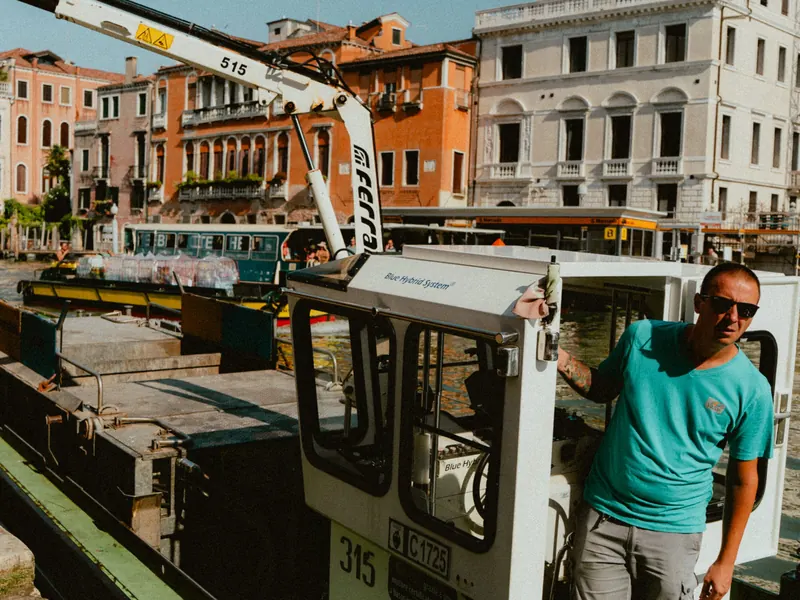 Un homme en t-shirt turquoise et lunettes de soleil, appuyé à la cabine d’une barge de travail équipée d’une grue, devant des palais aux façades ocre et rose et un arrêt de vaporetto.