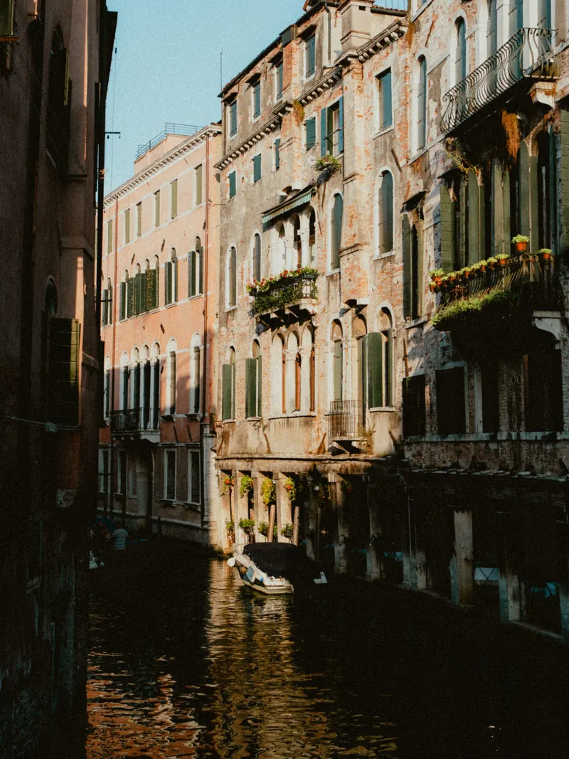Un canal étroit entre de hautes façades aux enduits usés, avec balcons et jardinières fleuries ; un bateau blanc est amarré dans l’ombre, le soleil éclaire le haut des maisons.