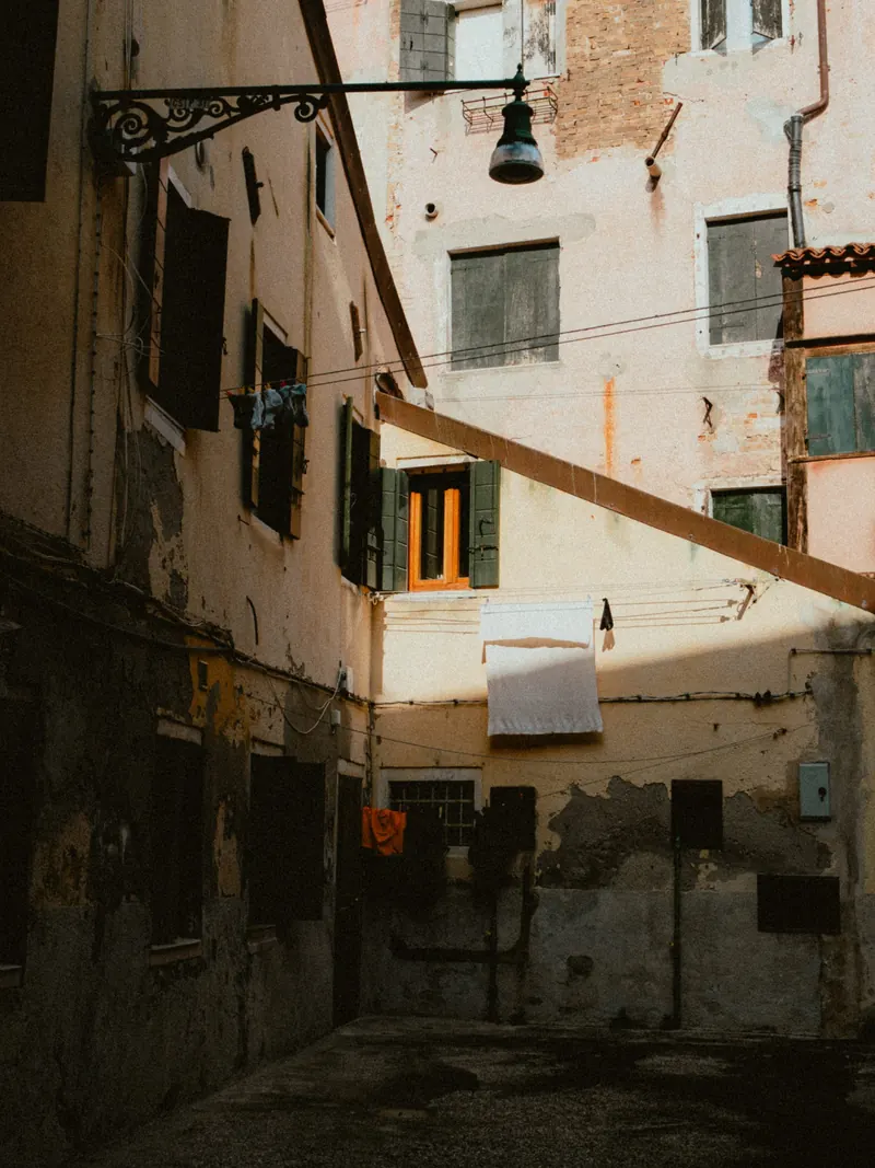 Une cour entre de hautes façades décrépies : du linge sèche aux fenêtres, une lanterne en fer forgé pend d’une console, une poutre de bois traverse la cour en diagonale ; seul le haut des murs est au soleil.