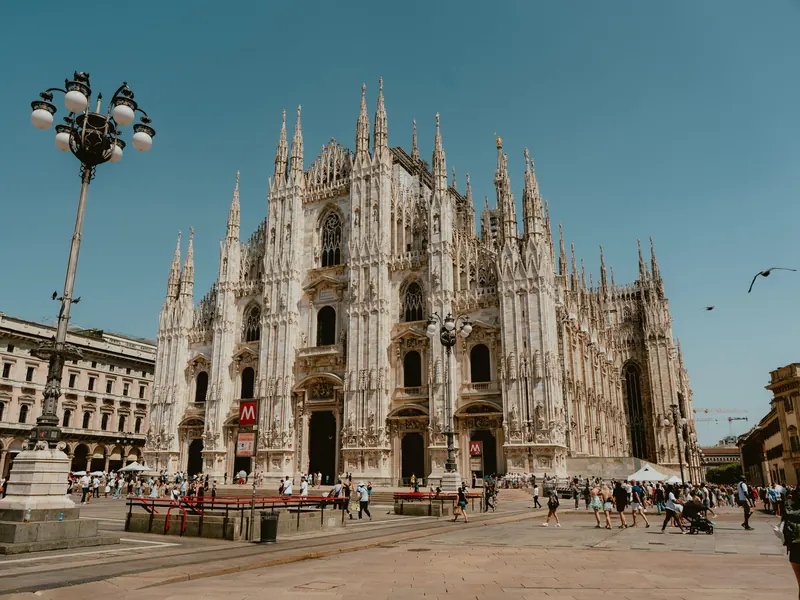 La façade gothique du Duomo de Milan et ses flèches, au bout d’une place où circulent des passants ; à gauche, un grand lampadaire à plusieurs globes, sous un ciel bleu.