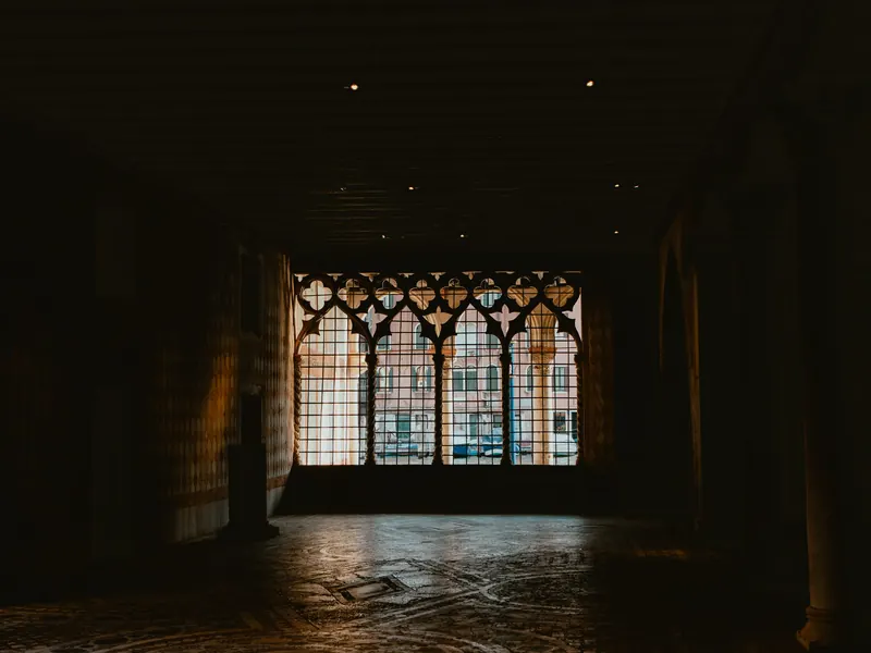 Dans la pénombre d’une grande salle, une fenêtre gothique à remplages, fermée d’une grille, s’ouvre à contre-jour sur des façades en plein soleil.