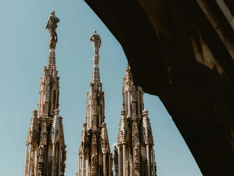 Trois flèches de marbre blanc surmontées de statues, sur un ciel bleu, encadrées par l’arc sombre d’une voûte.