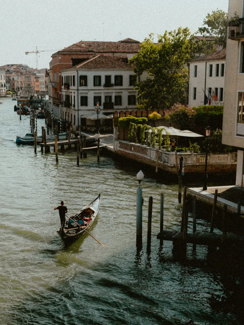 Un gondolier debout rame sur un large canal, entre des poteaux d’amarrage et un ponton ; au fond, des maisons aux toits de tuiles et un jardin en terrasse, dans la lumière du matin.