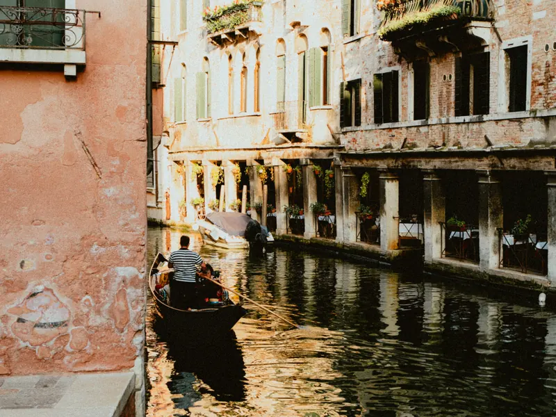 Un gondolier en marinière mène une gondole et ses passagers sur un canal étroit ; à droite, des tables dressées sous une galerie à piliers, à gauche un mur d’enduit rose écaillé, les façades dorées par le soleil.