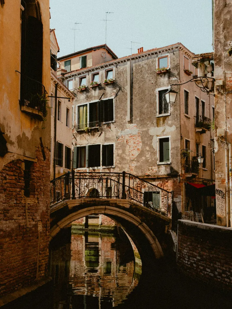 Un petit pont de pierre à rambarde de fer forgé enjambe un canal où son arche se reflète ; au-dessus, des façades à l’enduit écaillé et des jardinières aux fenêtres.
