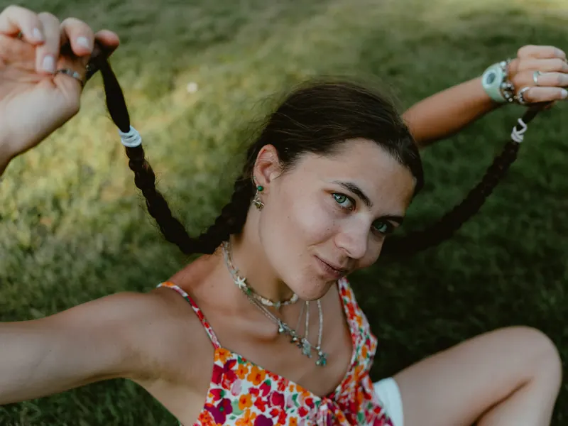 Portrait rapproché d’une jeune femme allongée dans l’herbe, qui tient écartées ses deux longues tresses brunes et regarde l’objectif.