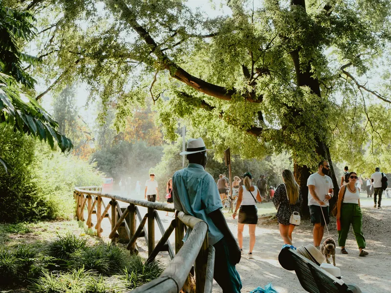 Des promeneurs, dont un avec un chien en laisse, sur une allée de terre sous de grands arbres ; au premier plan, un homme au chapeau blanc s’appuie à une barrière de bois.