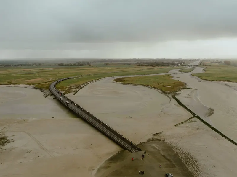 Vue plongeante depuis le Mont sur la baie : la passerelle et des promeneurs à son pied, le sable, les chenaux et les prés jusqu’à l’horizon, sous un ciel de pluie.