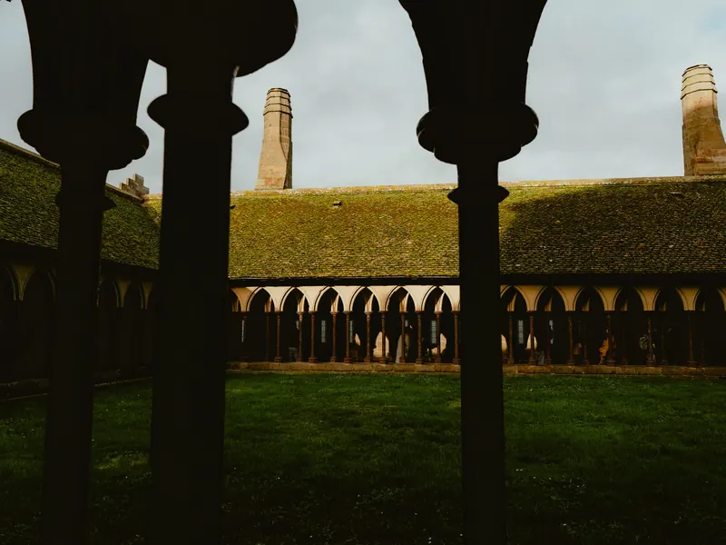 Le cloître de l’abbaye vu à contre-jour entre des colonnettes sombres : une pelouse, une galerie d’arcades et un toit couvert de mousse sous le ciel gris.