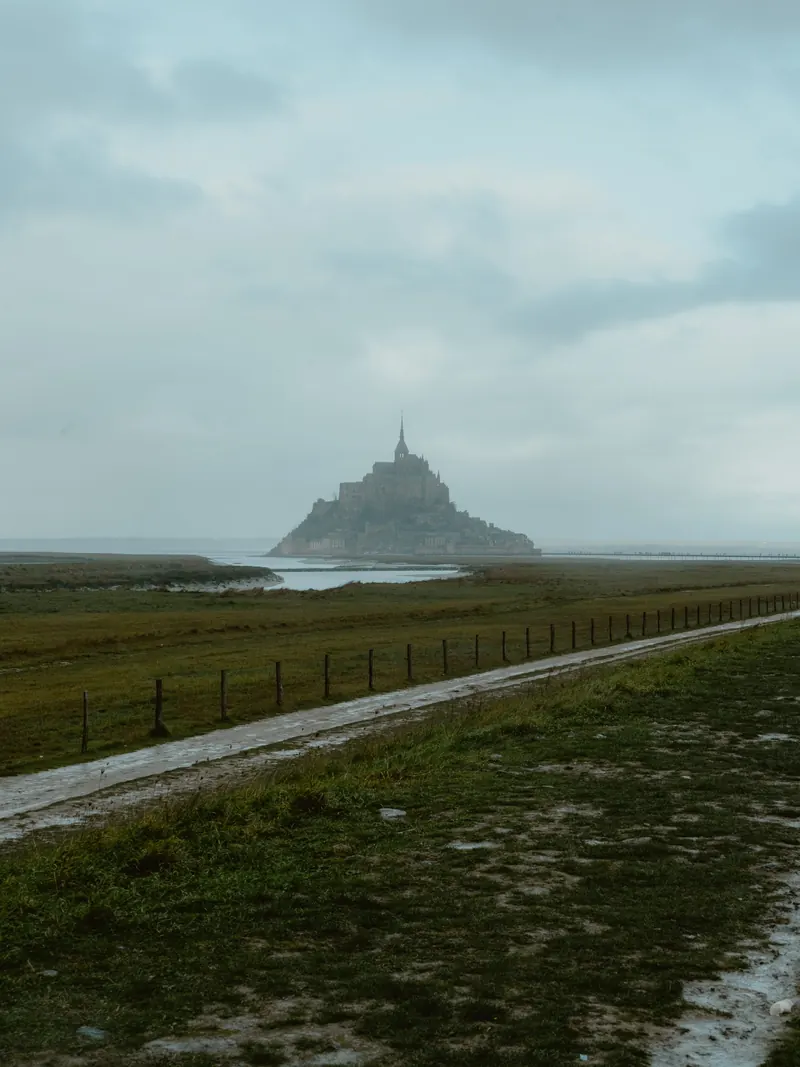 Le Mont-Saint-Michel au loin dans la brume, au bout d’un chemin détrempé longé d’une clôture et de prés.