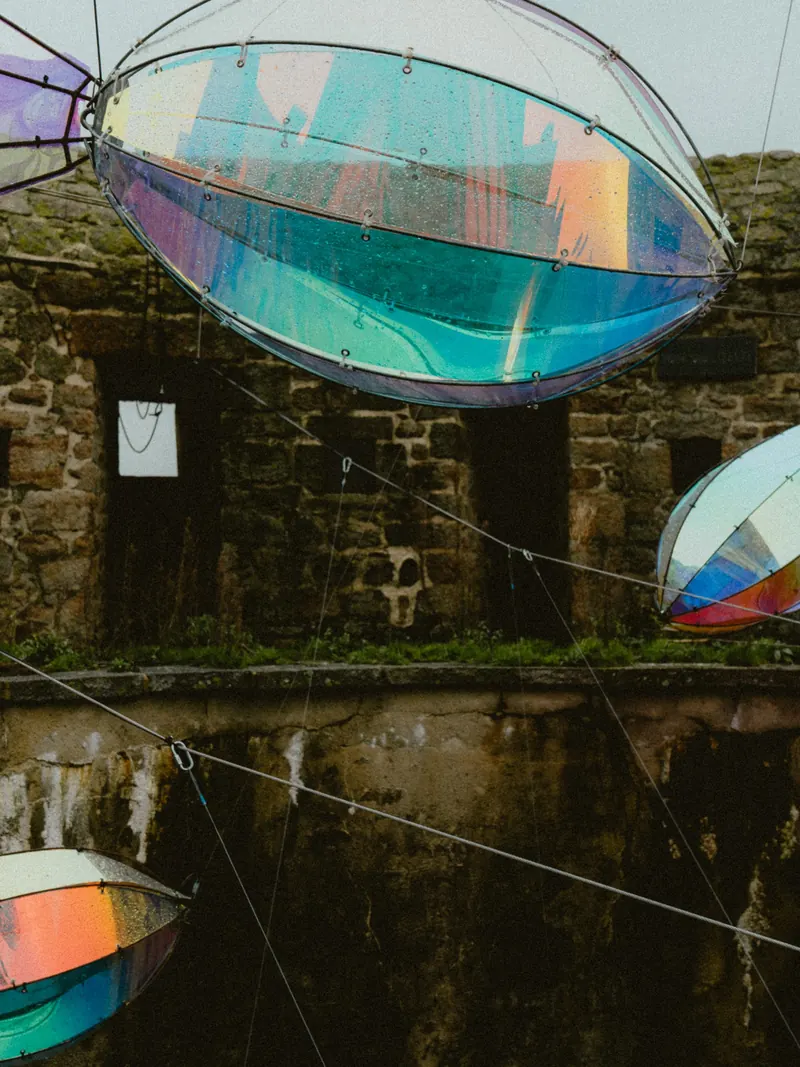 De grandes formes transparentes aux reflets irisés, suspendues à des câbles, flottent devant un mur de pierre couvert de mousse.